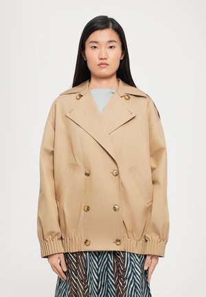 Giacca beige doppiopetto con bottoni dorati, vestibilità oversize, rever larghi e polsini elastici arricciati. Indossata sopra una gonna a fantasia.
