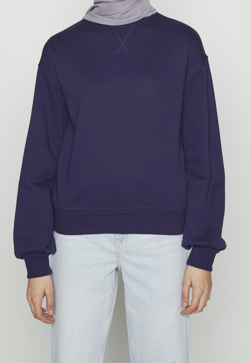 Personne portant un sweat-shirt bleu marine à manches longues et un jean bleu clair, avec un foulard gris clair visible au niveau du cou.