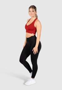 Sujetador deportivo rojo de canalé combinado con leggings negros de cintura alta. El modelo está de pie sobre un fondo simple, mostrando el ajuste y el diseño del conjunto.