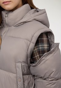 Gilet imbottito grigio chiaro con collo alto, chiusura con zip e tasche laterali; presenta un design trapuntato e testurizzato; indossato sopra una camicia a quadri.