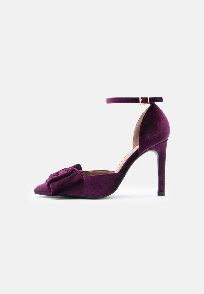 Sandal de salto alto em veludo roxo com tira no tornozelo, bico fino e detalhe decorativo de laço na parte da frente; textura suave e design elegante.