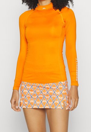 Vrouw draagt een aansluitend feloranje shirt met lange mouwen, gecombineerd met een oranje minirok met blauw-witte diamantvormen.