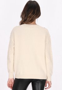 Crèmekleurige gebreide trui met een losse pasvorm, voorzien van textuur en kantpatronen op de mouwen en subtiele metallic accenten bij de schouders.