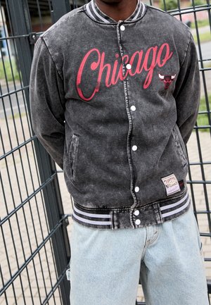 Person wearing a faded black Chicago Bulls varsity jacket with red lettering and light blue jeans standing against metal fence.
