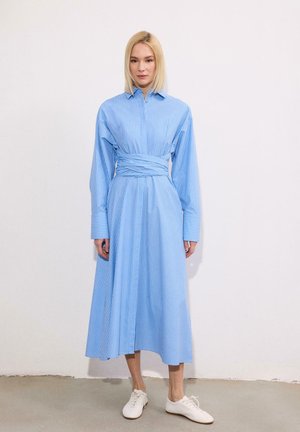 Γυναίκα με ίσια ξανθά μαλλιά που φοράει μπλε midi φόρεμα με λεπτές ρίγες και μακριά μανίκια, με ζώνη στη μέση, στέκεται σε τσιμεντένιο δάπεδο, με λευκά παπούτσια.