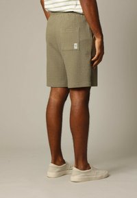 Olijfgroene shorts met een gestructureerd wafelpatroon, voorzien van een achterzak en een label. Getoond met lichtgekleurde instapschoenen.
