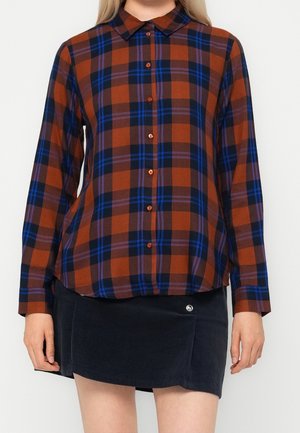 Woman wearing a long-sleeve rust and navy plaid button-up shirt with a black mini skirt, standing against a plain light background.