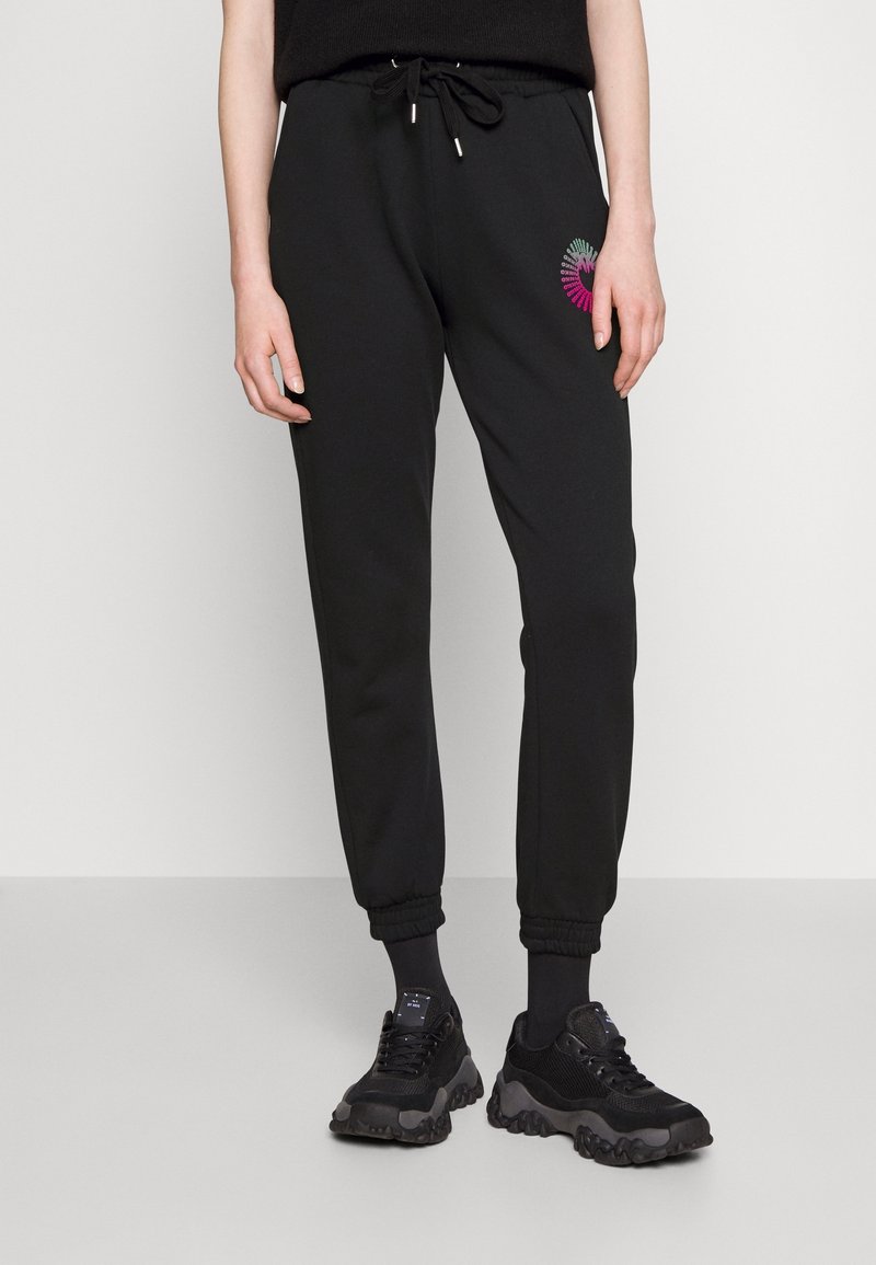 Czarne dresowe spodnie z elastycznymi mankietami, sznurkiem w talii oraz wielokolorowym okrągłym haftem na prawym udzie. Noszone z czarnymi sneakersami.