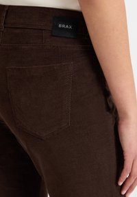 Bruine corduroy broek met een gestructureerde afwerking, voorzien van een achterzak en een zwart gelabelde tailleband gemarkeerd met "BRAX."