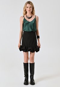 Camisola verde in satin con spalline sottili abbinata a una mini gonna nera con orlo arricciato. Stivali neri fino al ginocchio completano l'outfit.