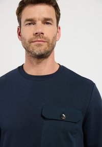 Marineblauwe sweatshirt van zacht materiaal, met een borstzak met knoop en een ronde hals. Gladde textuur en minimalistisch ontwerp.