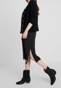 Svart texturerad cardigan med guldknappar, kombinerad med en svart knälång klänning med spetsdetaljer och sidofickor. Bärs med svarta ankelstövlar.