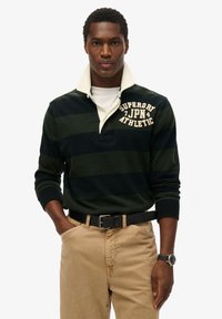 Ciemnozielona i granatowa koszulka rugby w paski z kremowym kołnierzykiem, z tekstem "Superdry JPN". Połączona z beżowymi spodniami i czarnym paskiem.