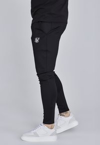 Čierne atletické jogger nohavice vyrobené z elastického materiálu, s prispôsobeným strihom, bočnými vreckami a bielym logom na stehne.