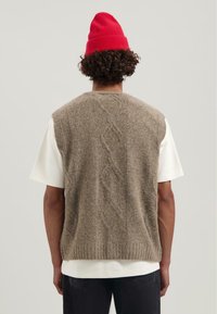Mann mit lockigem Haar, der eine rote Mütze, eine beigen Strickweste über einem weißen Kurzarmhemd und dunklen Jeans trägt, und mit dem Rücken zu einem einfarbigen Hintergrund steht.
