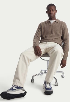 Mann sitzt auf einem drehbaren Bürostuhl, trägt einen braunen Pullover, weiße Hose und blau-graue Turnschuhe vor einem einfarbigen weißen Hintergrund.