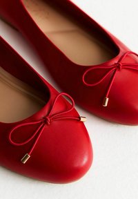 Ballerine in pelle rossa con punta arrotondata, caratterizzate da un fiocco decorativo e dettagli in tono oro su un materiale superiore liscio.