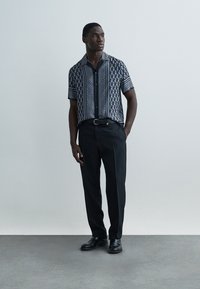 Short-sleeve button-up shirt in navy with geometric white patterns; paired with black trousers and black shoes, set against a neutral background.