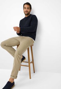 Marineblauer Strickpullover mit Ripptextur, beige schmale Hose und dunkle Sneakers auf einem Holzschemel vor einem schlichten weißen Hintergrund.