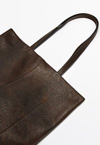 Bruine leren tote bag met een gestructureerd oppervlak, twee lange hengsels en een open bovenkant. Heeft een eenvoudig ontwerp en een minimalistische vorm.