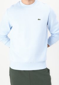 Man som bär en ljusblå sweatshirt med en liten grön krokodil-logotyp på bröstet och mörkgröna byxor, händerna bakom ryggen.