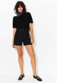 Pull noir à manches courtes associé à un short noir taille haute avec deux poches avant. Chaussures plates noires avec des accents cloutés.