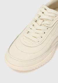 Valgekirju nahast toss, millel on tekstuurse tallaga, detailne õmblus ja lamedad kootud paelad, mis on seotud tavalise ristisõlmega.