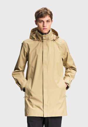 Jonge man draagt een beige regenjas met capuchon met handen in de zakken, staand tegen een effen lichtgrijze achtergrond.