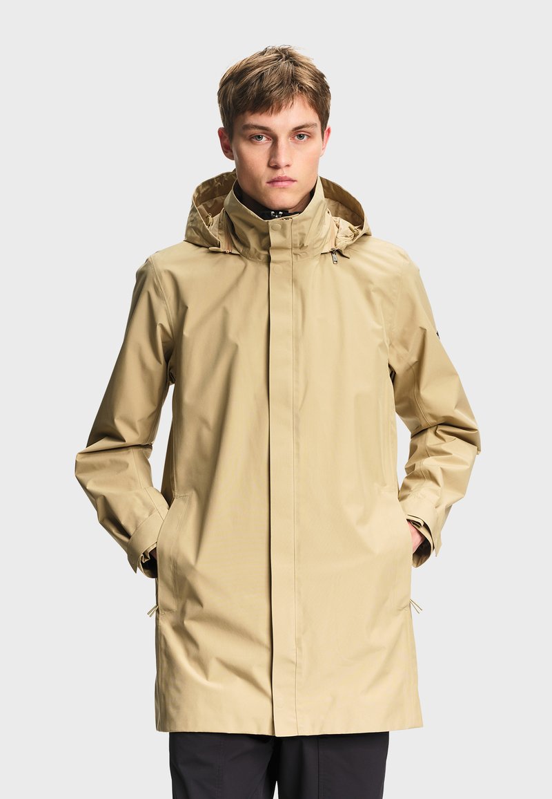 Jeune homme portant un imperméable à capuche beige avec les mains dans les poches, debout devant un fond uni gris clair.
