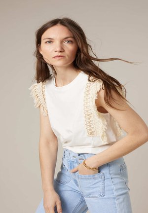 Femme aux longs cheveux bruns portant un haut blanc sans manches avec des détails en franges et un jean taille haute bleu clair, une main dans la poche.