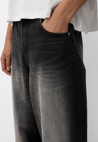 Osoba oblečená ve vybledlých černých džínách a bílém tričku, s pravou rukou položenou v přední kapse džínů.