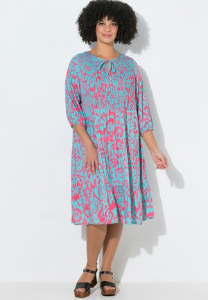 Frau mit lockigem Haar, die ein locker sitzendes, knieumspielendes Kleid in Türkis und Pink mit Muster, dreiviertellangen Ärmeln und schwarzen Sandalen mit Absatz trägt.