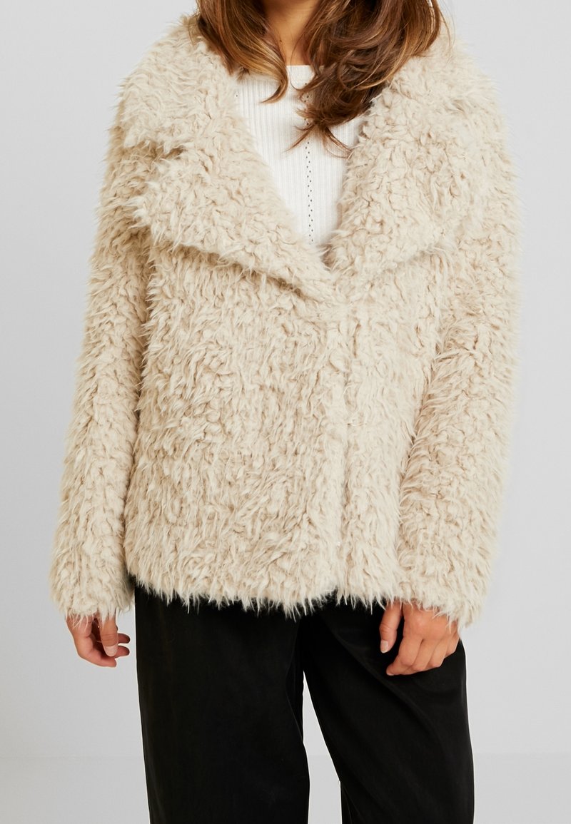 Femme portant un manteau beige duveteux texturé par-dessus un top blanc côtelé et un pantalon noir, debout devant un fond clair uni.