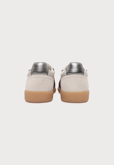 Paire de baskets vue de l’arrière, en daim beige clair, empiècements talon argentés et semelles en caoutchouc marron sur fond blanc.