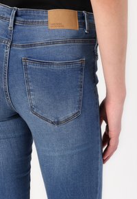 Jeans de mezclilla azul con corte slim, que cuentan con un bolsillo trasero y una etiqueta de marca de cuero marrón en la cinturilla. Textura suave, diseño clásico.