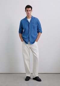 Camisa de mezclilla en azul claro con un bolsillo en el pecho, cierre de botones en la parte delantera y un cuello relajado, combinada con pantalones de pierna ancha de color crema y zapatos negros.