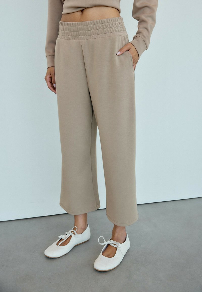 Personne portant un pantalon beige large coupé au-dessus de la cheville avec une taille élastique et des chaussures plates blanches à lacets, debout sur un sol gris.