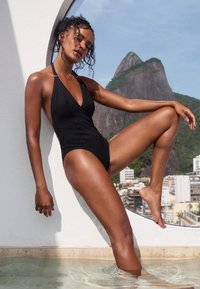 Black one-piece swimsuit with a deep V-neck, textured diamond pattern, and halter straps. Model poses with one leg in water against a white wall.