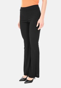 Donna che indossa pantaloni neri a campana aderenti, top arancione senza maniche e tacchi alti neri a punta, in piedi davanti a uno sfondo bianco.