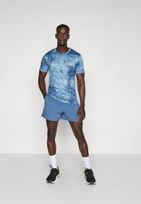Blauwe sportshirt met een tie-dye patroon en een logo, gecombineerd met effen blauwe shorts. Model draagt zwarte sneakers en witte sokken.