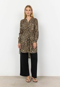 Lång skjorta med leopardmönster i lätt, genomskinlig tyg, knäppt framtill, knuten i midjan, tillsammans med svarta vida byxor och sandaletter med remmar.
