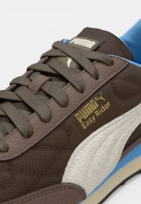 Gros plan sur une basket Puma Easy Rider marron avec des lacets marron foncé, une bande latérale blanche et une doublure intérieure bleue.