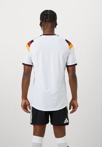 Männlicher Athlet mit geflochtenen Haaren, trägt ein weißes Fußballtrikot mit schwarzen, roten und gelben Schulternstreifen, schwarze Shorts und weiße Kniestrümpfe, von hinten gesehen.
