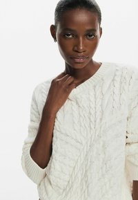 Maglione bianco a trecce con scollatura a costine e motivo testurizzato, con una leggera lucentezza e vestibilità rilassata. Il modello tiene la mano al collo.