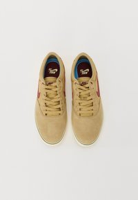 Hellbraune Wildledersneakers mit niedrigem Profil, die rote Akzente, beige Schnürsenkel und blaue Fütterung aufweisen. Flache Gummisohle mit cremefarbener Kante.