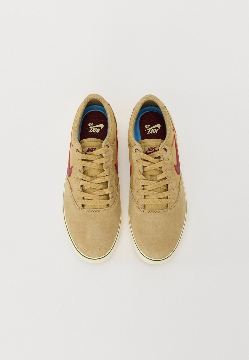 Zapatillas de ante en color marrón claro con un perfil bajo, que presentan acentos rojos, cordones beige y un forro azul. Suela plana de goma con borde en color crema.