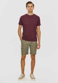 Burgundowa koszulka z krótkim rękawem połączona z oliwkowymi szortami; model stoi w jasnych sneakersach, minimalistyczny design w neutralnej kolorystyce.