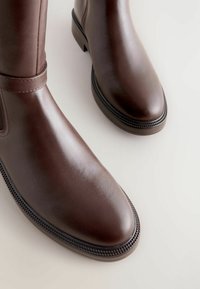 Botine maro din piele, cu vârf rotund, toc jos și cusături vizibile pe fundal alb.