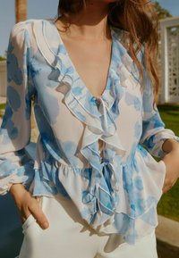 Lätt, genomskinlig blus med ett blått blommönster. Har volangkanter, en djup urringning och dragsnöre, kombinerad med vita byxor.