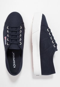 Námořnické modré plátěné tenisky s bílými gumovými podrážkami, kovovými očky, černými tkaničkami a viditelným logem Superga na jazyku a vnitřní podrážce.